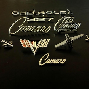 May include: A collection of vintage Chevrolet Camaro emblems and badges, including "Camaro" in script and block letters, "327", and a red, white, and silver emblem. The chrome-finished pieces are arranged on a dark surface.