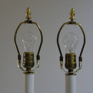 May include: Two white table lamps with brass accents and clear light bulbs. The lamps have a classic design, featuring a gold finial at the top and a curved brass support for the bulb. The lamps are suitable for a bedroom or living room.