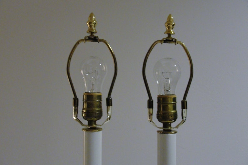 May include: Two white table lamps with brass accents and clear light bulbs. The lamps have a classic design, featuring a gold finial at the top and a curved brass support for the bulb. The lamps are suitable for a bedroom or living room.