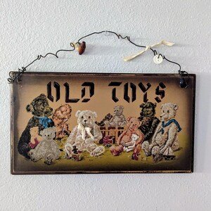 May include: A rectangular wooden sign with a dark brown border and a tan center featuring the text OLD TOYS in black. The illustration depicts several teddy bears in various shades of brown, tan, and gray, some wearing ribbons or scarves.