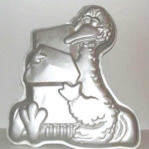 Puede incluir: Un molde para pasteles de color plateado con forma de pájaro de dibujos animados, posiblemente un pato o un ganso, sosteniendo un objeto rectangular. El molde tiene detalles, incluyendo ojos, pico y plumas. El molde es de metal.