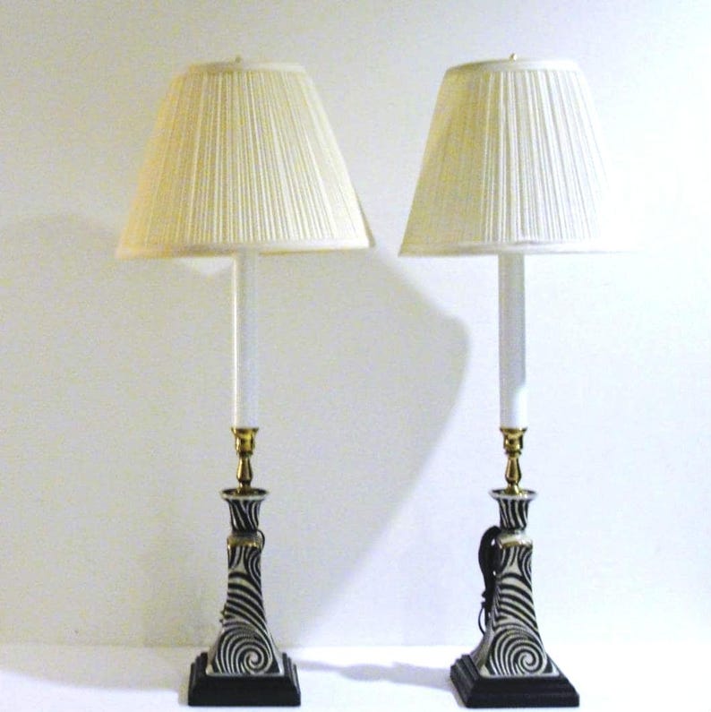 May include: Pair of decorative table lamps. Each lamp features a cream-colored pleated shade, a white column, and a base with a black and white swirl design. The lamps have a brass-colored accent and a square black base.