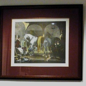 Puede incluir: Impresión enmarcada de una escena de establo con caballos y figuras. La obra de arte presenta un caballo blanco con una manta amarilla, rodeado de personas con ropa de época. El marco es marrón oscuro con un paspartú granate.