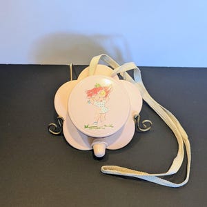 May include: A vintage pink compact mirror with a floral design. The compact has a round mirror with a cartoon girl illustration. It features a cream-colored strap and decorative metal accents.