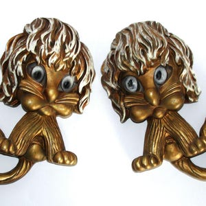 Puede incluir: Dos adornos de pared vintage con forma de gato. Cada uno tiene un cuerpo dorado con detalles de pelaje blanco y dorado. Los gatos tienen ojos grandes y expresivos y están sentados con la cola enroscada.