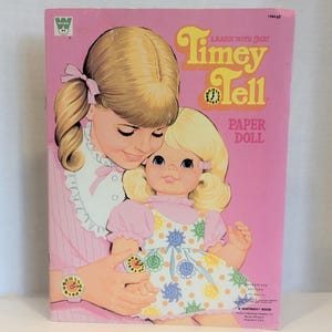 May include: Vintage "Timey Tell" paper doll book. The cover features an illustration of a girl holding a doll, both with blonde hair and wearing pink. The book's title is in orange and yellow, with the words "Learn with Me!" above it.