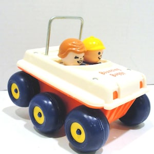 Puede incluir: Coche de juguete vintage con dos figuras sonrientes. El coche tiene una parte superior blanca, una base naranja y seis ruedas azul marino con centros amarillos. El texto "Bouncing Buggy" es visible. Una barra de metal plateado sirve como barra antivuelco.