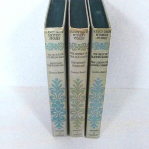 May include: Three vintage Nancy Drew Mystery Stories books. The book spines are off-white with teal and green floral designs. Titles include "The Clue in the Crumbling Wall," "The Secret of the Old Clock," and "The Ghost of Blackwood Hall."
