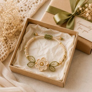 May include: A beige macrame bracelet featuring a central flower motif with six petals and two green thread-embroidered leaves on each side. The bracelet includes small gold-toned beads and an adjustable sliding knot closure, presented in a box.