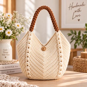 Könnte beinhalten: Eine cremefarbene Häkeltasche mit V-förmiger Öffnung, einem einzelnen geflochtenen braunen Henkel und einem zentralen Holzknopfverschluss. Die Tasche ist mit einem strukturierten Fischgrätmuster und vertikalen Reihen aus Noppenstichen versehen.