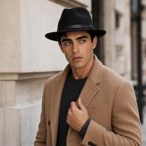 Op de afbeelding: Een zwarte vilten fedora-hoed met een dunne zwarte leren band rond de kroon, gedragen met een donker antracietgrijze ribgebreide trui en een beige wollen overjas met inkepingen in de revers en donkere knopen op de voorzijde en mouwboorden.