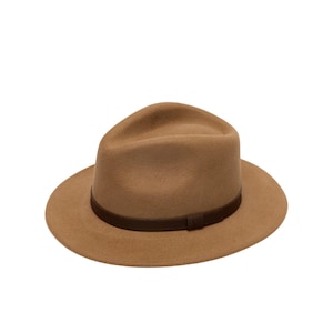 Op de afbeelding: Een beige fedora hoed met een gestructureerde kroon en een brede rand. De hoed is voorzien van een donkerbruine band rond de basis van de kroon, wat een duidelijk contrast vormt met de lichte beige kleur van het viltmateriaal van de hoed.