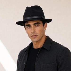 Op de afbeelding: Een zwarte fedora hoed met een gladde vilttextuur en een dunne zwarte leren band rond de kroon. De hoed heeft een gestructureerde rand en een ingeknepen kroonontwerp, wat zorgt voor een klassiek silhouet boven een donkergrijze jas.