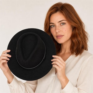 Könnte beinhalten: Ein einfarbiger schwarzer Fedora-Hut mit breiter Krempe und einem dünnen, passenden schwarzen Band um die Krone. Der Hut zeichnet sich durch ein strukturiertes Design mit eingekniffener Oberseite und eine glatte, matte Oberfläche aus.