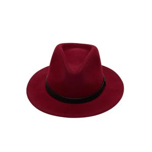 May include: A deep red fedora hat featuring a structured crown with a central crease and a wide, flat brim. A thin, smooth black band encircles the base of the crown, providing a distinct contrast against the solid red felt-like material of the hat.