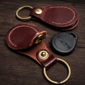 May include: Two dark brown leather key fobs with brass-colored metal snap closures and circular key rings. One fob is closed, while the other is open, revealing a small black circular electronic key tag with the text MF2.0 embossed on its surface.