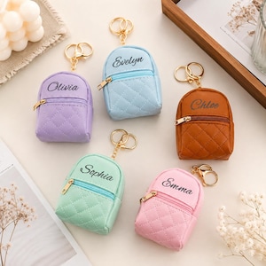 May include: Five miniature quilted backpack keychains in lavender, light blue, brown, mint green, and pink. Each features a gold-tone zipper, a gold-tone keyring, and a custom name embroidered in black script: Olivia, Evelyn, Chloe, Sophia, and Emma.