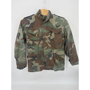 May include: A camouflage field jacket featuring a woodland pattern in shades of green, brown, and black. The jacket includes a high collar, long sleeves with buttoned cuffs, shoulder epaulets, and four front pockets with buttoned flaps.