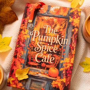 Op de afbeelding: Een hardcover boek getiteld The Pumpkin Spice Cafe met een omslagillustratie van een winkelpui omringd door levendige oranje en gele herfstbladeren, voorzien van een rood lint als bladwijzer met een zilveren hartvormige bedel.