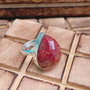 May include: A silver ring featuring a large, faceted, pear-shaped red gemstone set in a smooth metal bezel. The stone displays deep crimson hues with natural internal inclusions and a polished surface, mounted on a simple, polished silver band.