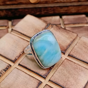 May include: A silver ring featuring a large, rectangular, polished light blue stone with white marbled veining. The stone is set in a smooth silver bezel with a simple, polished silver band that has a split design on the sides of the setting.