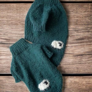Puede incluir: Dos suéteres de punto color verde azulado oscuro para mascotas, cada uno con cuello alto acanalado y mangas cortas. Cada suéter está decorado con un pequeño motivo de oveja blanca y esponjosa con detalles negros en el dobladillo.