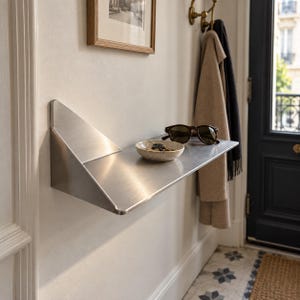 May include: A brushed metal wall-mounted shelf with a triangular side support. The flat surface holds a small, round, off-white ceramic bowl containing metal keys and a pair of dark-framed sunglasses with brown lenses, positioned against a white wall.