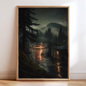 May include: A framed wall art print depicting a dark, rainy night scene in a small town. The image features a wet road reflecting warm orange lights from a building, surrounded by tall pine trees, misty mountains, and a moody, overcast sky.