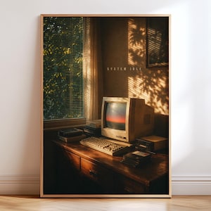 May include: A framed wall art print featuring a vintage beige computer monitor and keyboard on a wooden desk. The screen displays a sunset gradient and the text SYSTEM IDLE. Sunlight filters through window blinds, casting shadows on the wall.