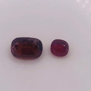Puede incluir: Dos piedras preciosas sueltas con talla cojín. La piedra más grande a la izquierda es de un color púrpura rojizo profundo, mientras que la más pequeña a la derecha es de un rojo vibrante y saturado. Ambas tienen superficies facetadas.