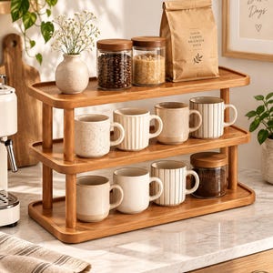 Könnte beinhalten: Ein zweistöckiges Küchenregal aus Holz mit verschiedenen Keramiktassen, Glasbehältern mit Holzdeckeln für Kaffeebohnen und Zucker, einer kleinen Vase mit weißen Blumen und einer braunen Papiertüte mit der Aufschrift Good Coffee Brighter Days.