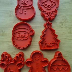 Pode incluir: Um conjunto de sete cortadores de biscoitos de plástico vermelho com formas temáticas de feriado, incluindo um boneco de neve, uma pessoa de óculos, o Pai Natal, uma árvore de Natal com presentes, uma rena, um boneco de gengibre e uma bola.