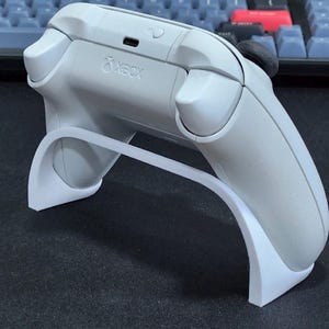 May include: A white wireless game controller resting on a white curved plastic stand. The controller features the Xbox logo, a USB-C charging port, and a pairing button on the top edge. The device is matte white with black thumbsticks visible.