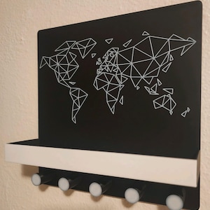Può includere: Un organizer da parete nero con il contorno di una mappa del mondo geometrica in linee bianche. L'unità include una mensola orizzontale bianca e cinque ganci cilindrici neri con estremità circolari bianche per appendere oggetti.