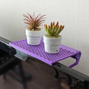 Puede incluir: Un estante de plástico morado con un patrón de rejilla hexagonal montado sobre una pantalla de televisión negra. Dos pequeñas macetas blancas con plantas artificiales están colocadas sobre el estante, una con follaje rojo y otra con hojas verdes.
