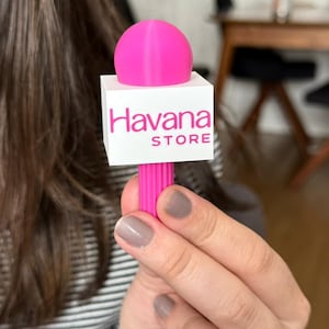 May include: A bright pink microphone prop featuring a white square flag with the text Havana STORE printed in pink. The microphone has a rounded top and a vertically ribbed handle, designed as a handheld accessory for branding or promotional use.