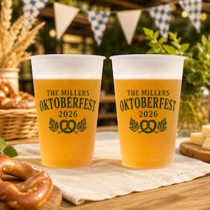 May include: Two frosted plastic cups filled with golden beer, each featuring dark green text that reads THE MILLERS OKTOBERFEST 2026 above a graphic of a pretzel flanked by two hop cones. The cups are positioned side by side on a light fabric surface.