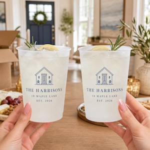 May include: Two frosted plastic cups filled with ice, a lemon slice, and a sprig of rosemary. Each cup features a blue graphic of a house and the text: THE HARRISONS, 18 MAPLE LANE, EST. 2026. The cups are held up against a blurred indoor background.