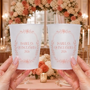 Op de afbeelding: Twee bekers van mat plastic met een bloemrijk, ovaal frame in roségoud. De tekst in het midden luidt "ISABELA'S QUINCEAÑERA 2026" in een schreeflettertype. De bekers zijn doorschijnend met een gladde afwerking en een eenvoudige rand.