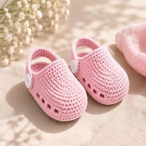 May include: A pair of light pink crocheted baby shoes featuring a rounded toe, a heel strap, and a single white button on each side. The shoes have small rectangular cutouts along the sides and are crafted from a textured yarn material.