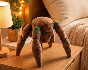 May include: A plush toy shaped like a rock creature with a brown, cracked, stone-like texture and small, bright green patches scattered across its rounded body and four long, sturdy limbs that support its weight on a wooden surface.
