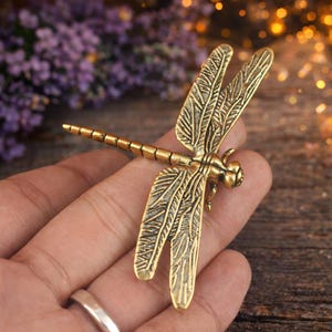 May include: A metallic gold-toned dragonfly brooch featuring intricate wing detailing and a segmented body. The dragonfly has four extended wings with fine linear patterns and a textured surface, showcasing a realistic insect design.