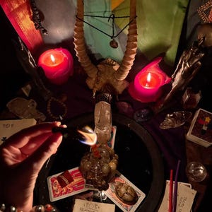 May include: A ritualistic arrangement featuring a pair of curved animal horns, two lit pink candles, a clear quartz crystal, a stone scarab, and tarot cards. A hand holds a burning incense stick over a paper with the text ABRACADABRA written in a pyramid.