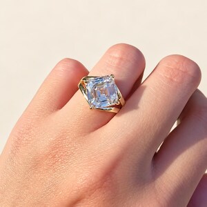 Peut inclure: Une bague en or avec une grande pierre transparente de taille carrée et à facettes étagées. L'anneau est en or jaune poli et présente un sertissage de style bypass qui maintient la pierre centrale au niveau des coins.