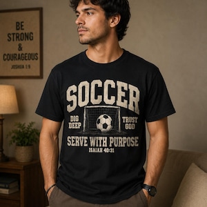 Op de afbeelding: Een zwart t-shirt met korte mouwen en een afbeelding van een voetbal in een doelnet. De tekst SOCCER, DIG DEEP, TRUST GOD, SERVE WITH PURPOSE en ISAIAH 40:31 staat in een verweerd, gebroken wit lettertype op de borst van het kledingstuk.