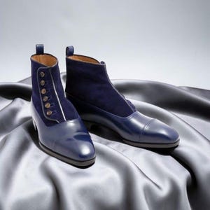 Könnte beinhalten: Ein Paar marineblaue Stiefeletten mit einer glatten Lederkappe und einem Obermaterial aus Wildleder. Die Stiefel verfügen über einen seitlichen Verschluss mit einer vertikalen Reihe aus fünf kleinen runden Knöpfen und einer Zuglasche.
