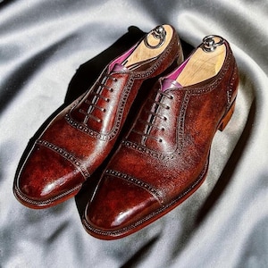 May include: A pair of dark brown leather oxford shoes featuring a cap toe with decorative brogue detailing, thin dark brown laces, and a polished finish. Each shoe includes a wooden shoe tree with a metal ring handle and a purple inner lining.