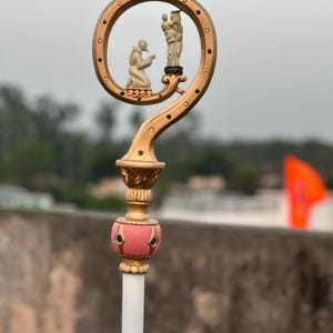 May include: A gold-toned metal crozier featuring a curved top with a circular opening. Inside the circle are two small off-white figures, one kneeling and one standing. Below the circle is a pink spherical accent with green inlays on a white staff.