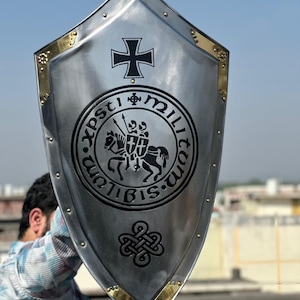 May include: A silver-colored heater shield featuring gold-toned metal trim along the edges with decorative rivets. The center displays a black iron cross, a circular emblem depicting two knights on a horse with the text Sigillum Militum Xpsti, and a knot.