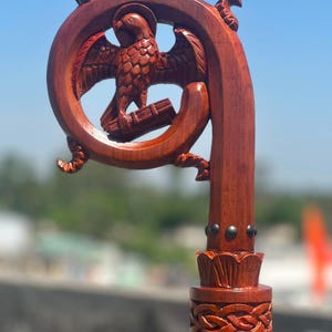 May include: A carved wooden crozier featuring a circular top with an eagle perched on a book. The wood has a reddish-brown finish. Below the circular head, the staff includes a decorative band with an intricate braided pattern and metal studs.